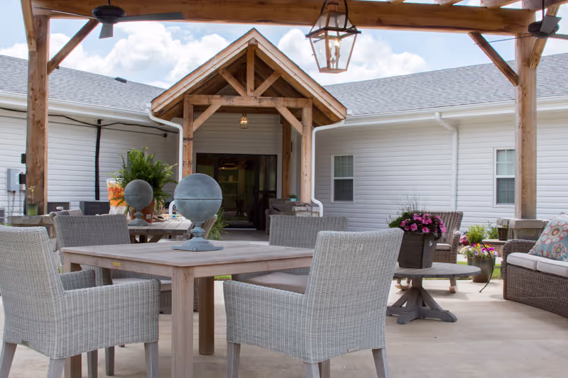 Covered outdoor patio with wooden pergola, wicker chairs and tables arranged in front of a single-story building entrance.