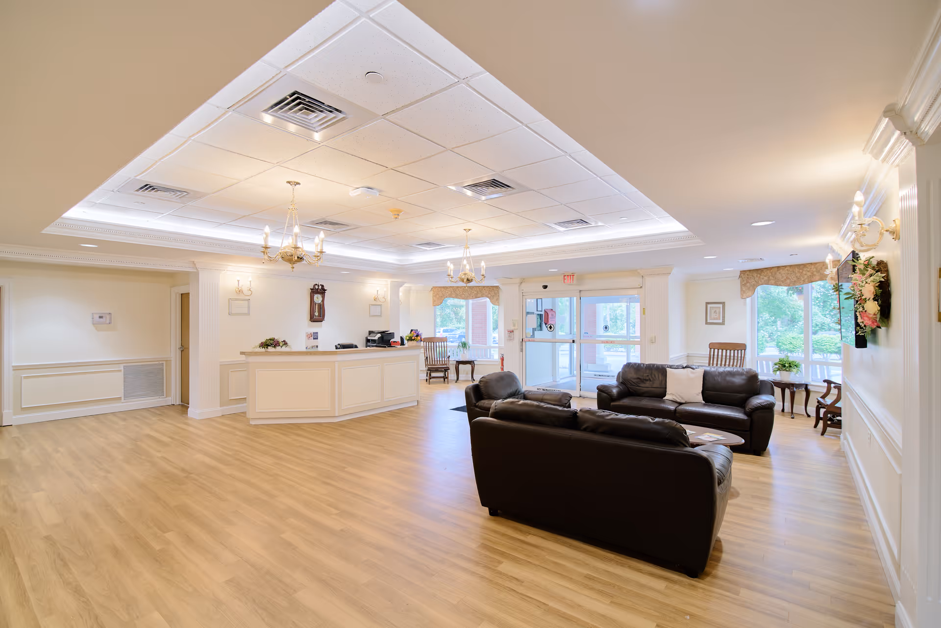 A spacious and well-lit senior living facility lobby with light wood flooring, white walls, and a white reception desk. There are two dark leather sofas arranged around a small round coffee table. The ceiling features recessed lighting and chandeliers. Large windows and glass doors allow natural light to enter, and there are decorative wall sconces and a clock on the wall behind the reception desk.