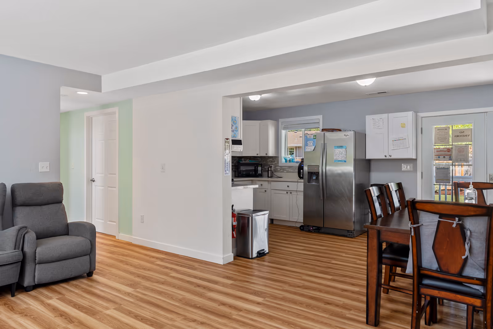 Interior view of a senior living facility showing a living area with two gray recliner chairs on the left, a wooden dining table with chairs on the right, and a kitchen area in the background with white cabinets, a stainless steel refrigerator, and a window. The floor is wood laminate, and there is a door with glass panels leading outside.