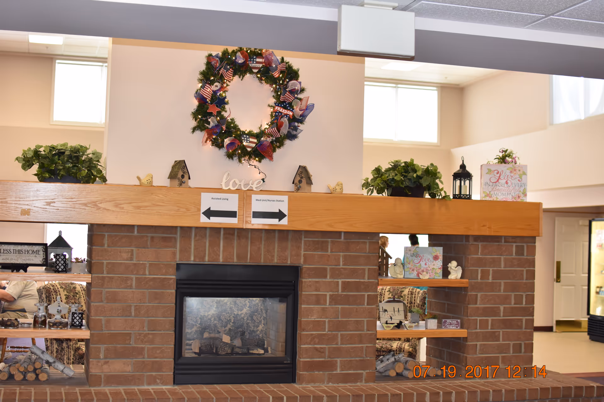Indoor common area with a brick fireplace topped by a decorated wreath and shelves holding plants and decor.