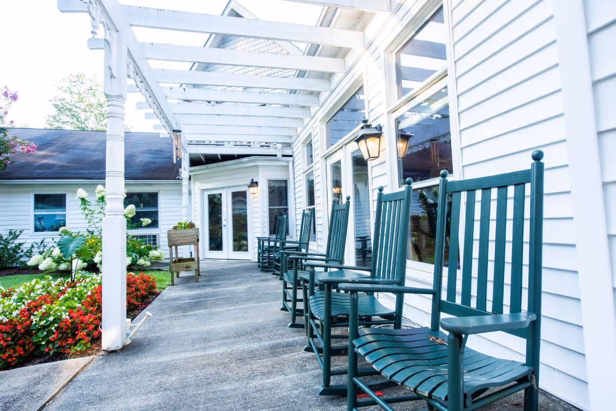 Covered outdoor patio at a senior living facility with a row of green rocking chairs along a white-sided building and flowerbeds.