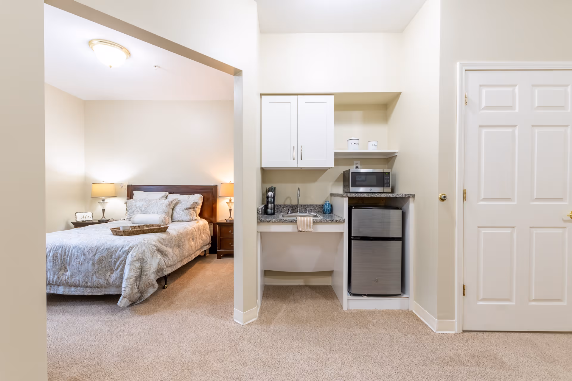 A cozy bedroom with a neatly made bed featuring patterned bedding and multiple pillows, flanked by two bedside tables each with a lamp. Adjacent to the bedroom is a small kitchenette area with a sink, a mini refrigerator, a microwave, and white cabinets. The walls are painted a light beige color and the floor is carpeted.