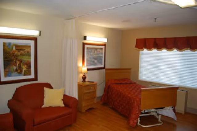 A cozy senior living bedroom with a single bed covered in a red patterned bedspread, a wooden nightstand with a lamp, a red armchair with a yellow pillow, two framed paintings on the walls, a large window with blinds and a red valance, and a privacy curtain partially drawn.