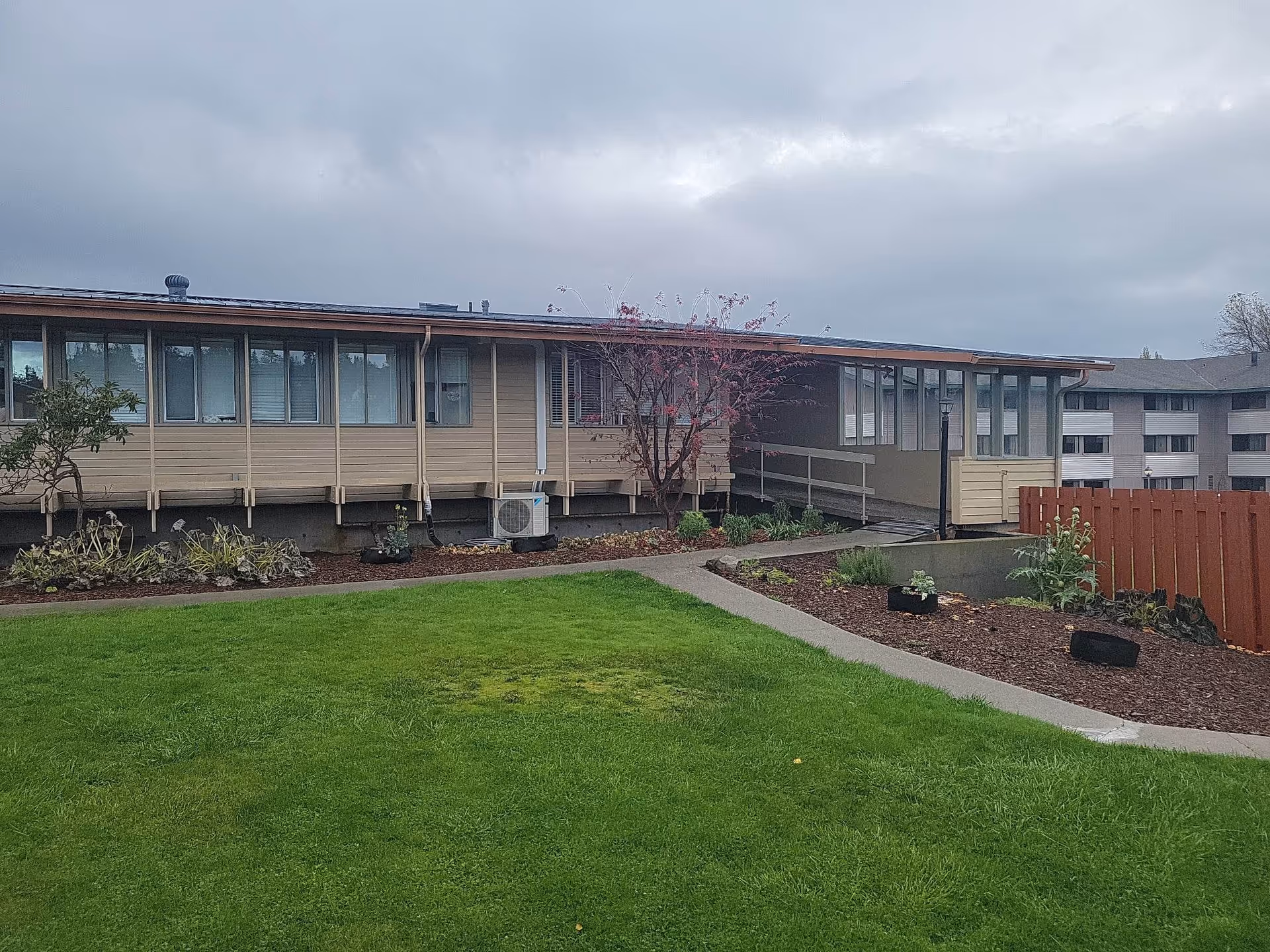 Exterior view of a single-story building with beige siding and multiple windows, surrounded by a green lawn and landscaped garden beds. A concrete pathway leads to a covered entrance with a ramp. The sky is overcast.
