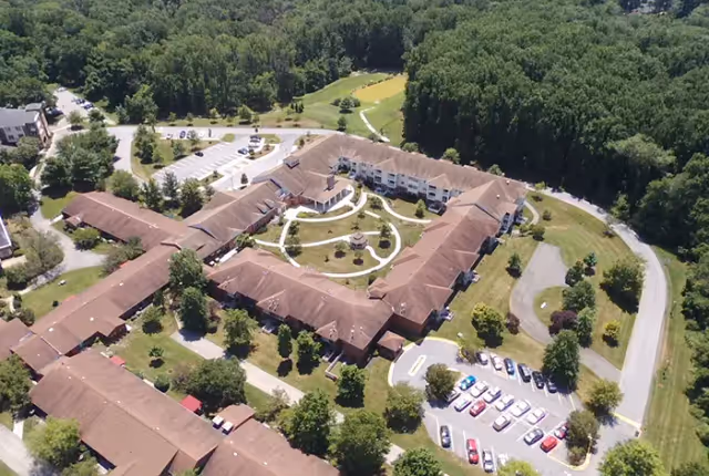 Aerial view of Resorts of Augsburg senior living facility showing a large building complex with a central courtyard featuring walking paths and a gazebo, surrounded by trees and parking lots with several cars.