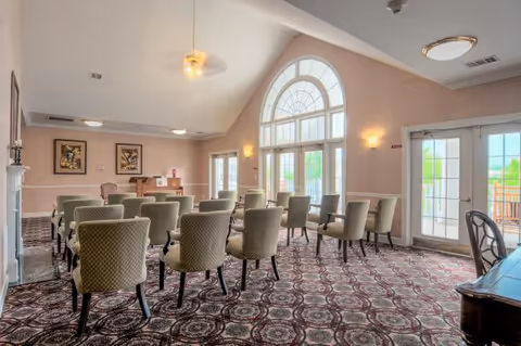 A well-lit room with rows of upholstered chairs arranged facing a piano and a fireplace. The room features large arched windows and glass doors allowing natural light to enter, with patterned carpet flooring and framed artwork on the walls.