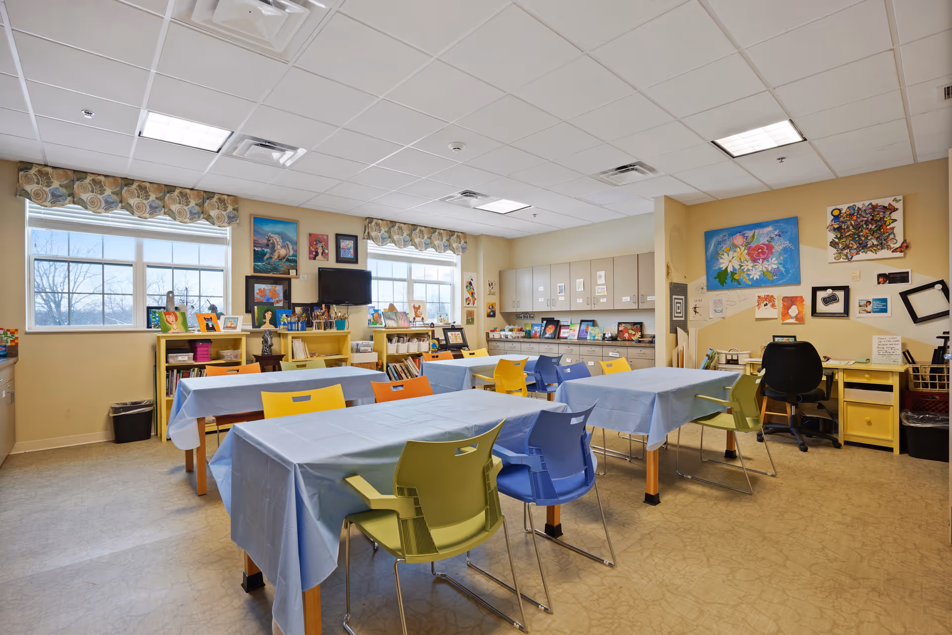 A bright and colorful activity room with several tables covered in light blue tablecloths and surrounded by multicolored chairs. The room has large windows with floral valances letting in natural light. The walls are decorated with various paintings and artwork. There are shelves filled with books, art supplies, and framed pictures. A black office chair is positioned at a yellow desk in the corner.
