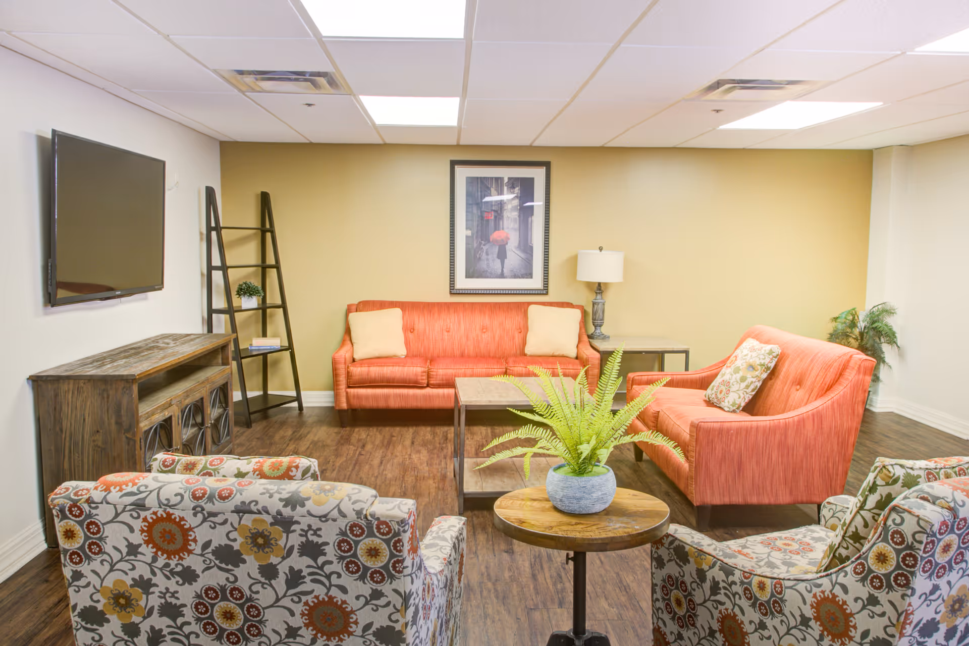A cozy living room with two coral-colored sofas, two floral-patterned armchairs, a wooden coffee table, and a small round side table with a potted fern. A flat-screen TV is mounted on the wall above a rustic wooden media console. A framed picture hangs on the beige wall behind the sofas, and a floor lamp is placed next to one of the sofas. The room has wood flooring and a white ceiling with recessed lighting.