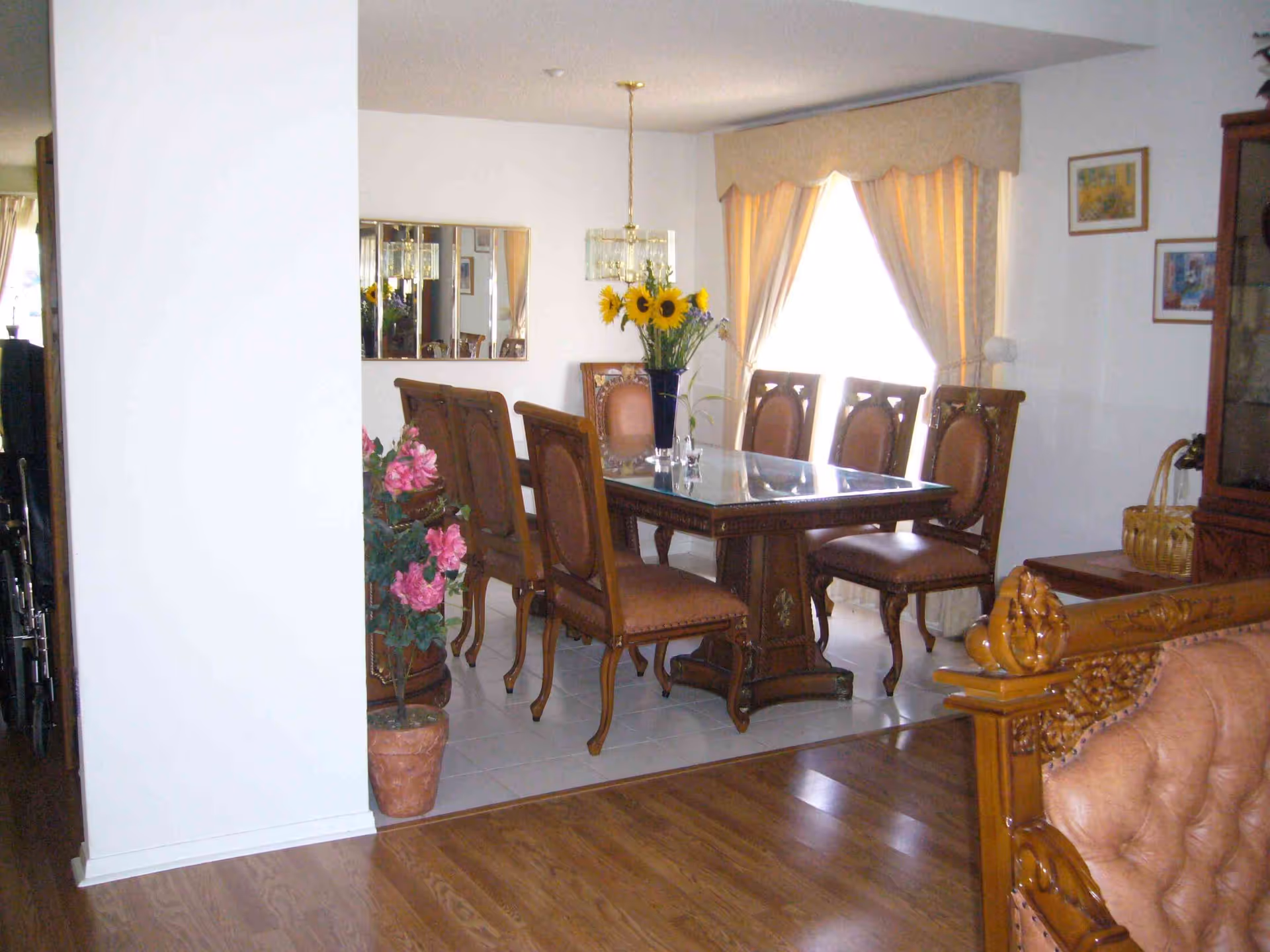 A dining room with a wooden dining table and six matching chairs with upholstered seats. The table has a glass top and a vase with sunflowers in the center. There is a large window with curtains allowing natural light to enter the room. A mirror hangs on the wall, and there are framed pictures on the walls. A potted plant with pink flowers is placed near a white wall, and part of a wooden sofa with carved details is visible in the foreground.