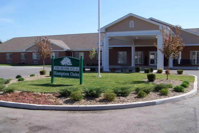 Front exterior view of Hampton Oaks Health Campus building with a green sign in the landscaped front yard that reads 'The Residence at Hampton Oaks'. The building has a covered entrance with white columns and a well-maintained lawn with small trees and shrubs.