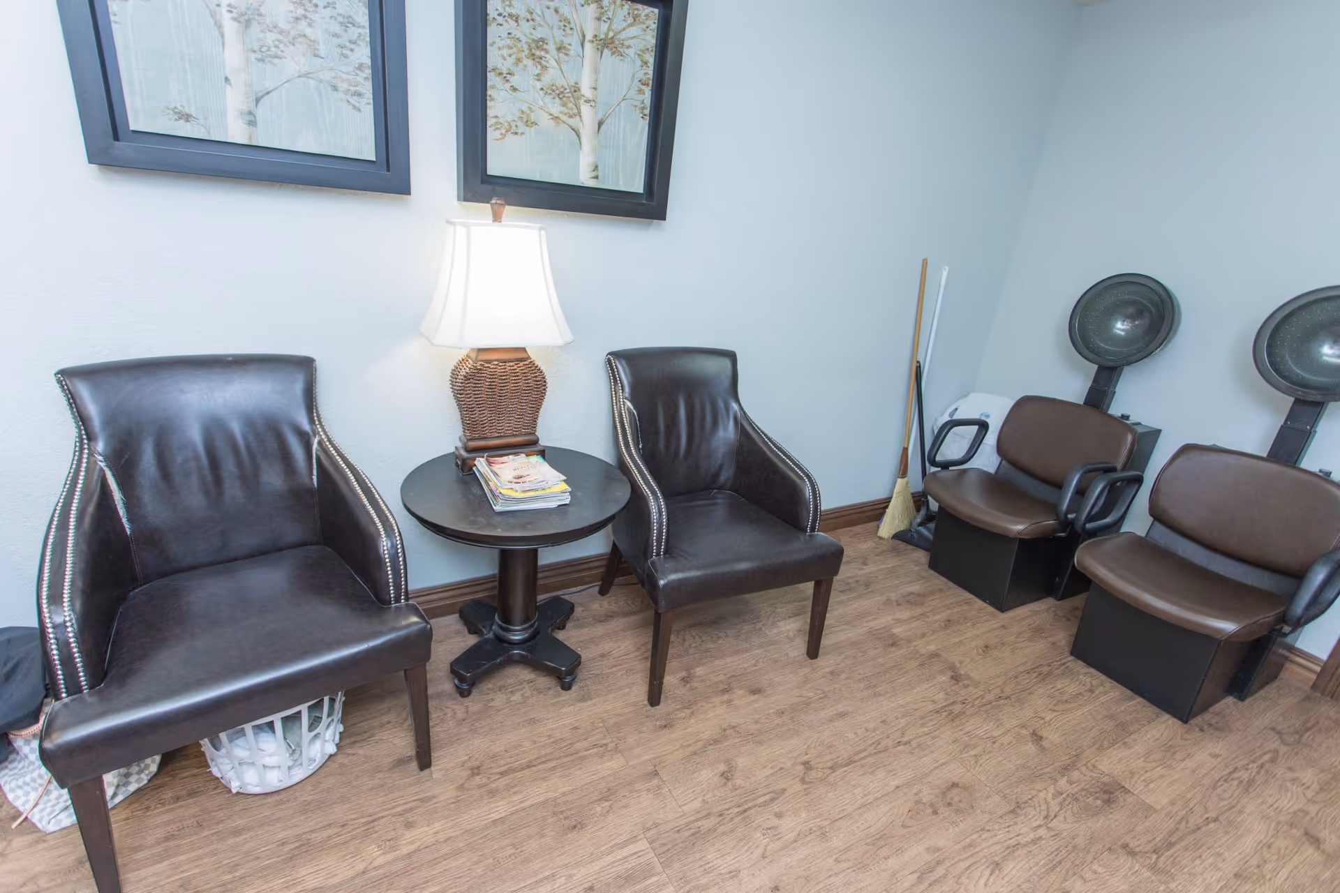 A small room with two black leather chairs separated by a round table with a lamp and magazines on it. On the right side, there are two brown salon hair dryer chairs with hooded dryers. The floor is wooden, and two framed pictures of trees hang on the light blue wall.