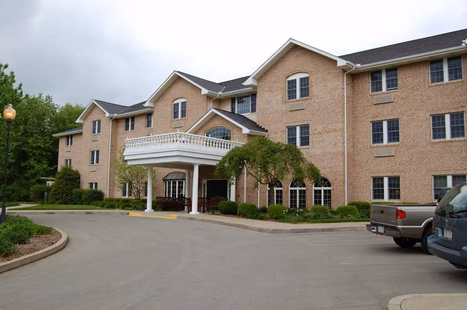 Exterior view of a three-story brick building with multiple windows, a covered entrance with white columns, landscaped greenery, and a curved driveway with parked vehicles.