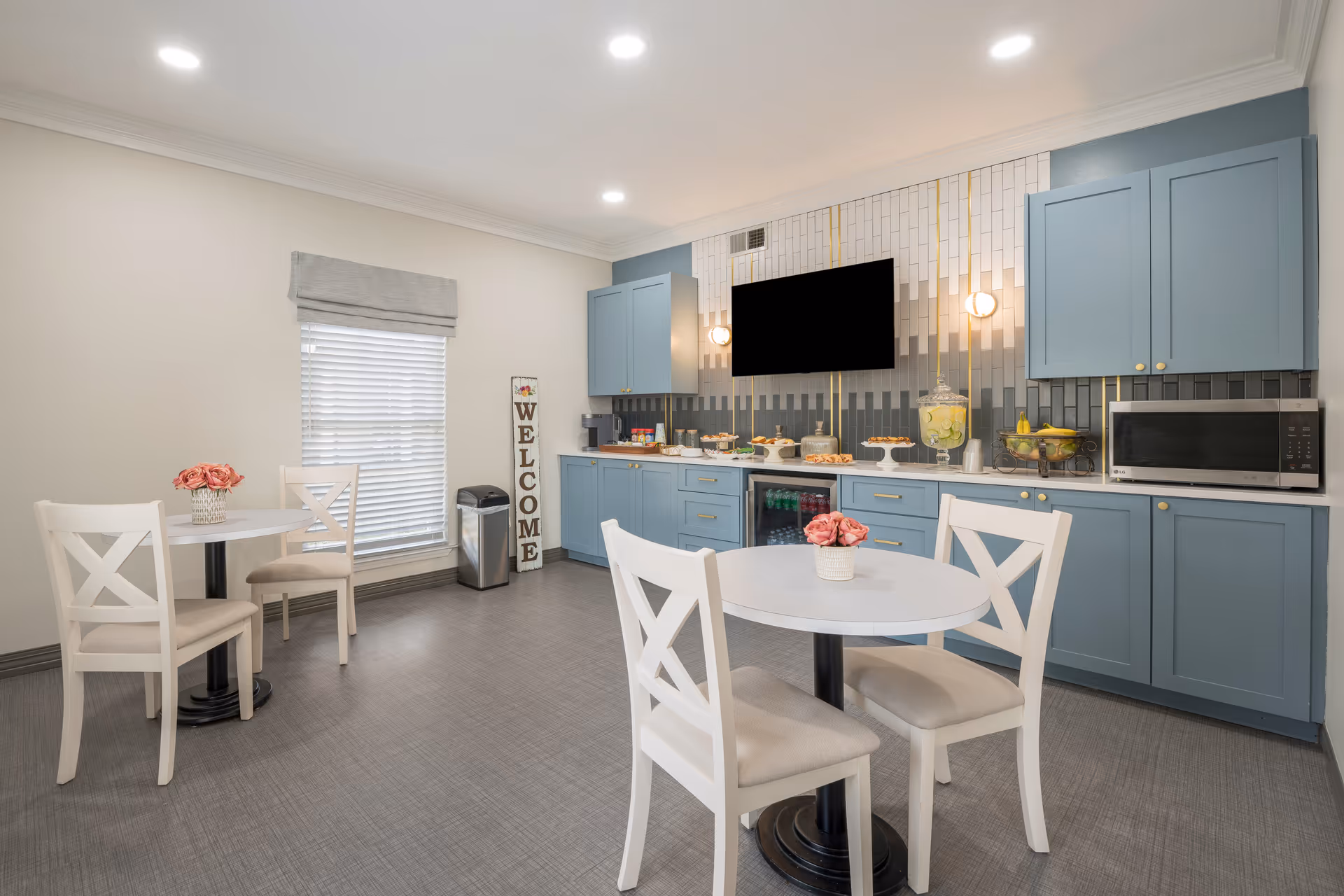 A bright and clean dining area with two small round tables, each with two white chairs. The tables have small flower arrangements. The back wall features blue cabinets, a microwave, a small refrigerator, and a countertop with various snacks and a beverage dispenser. A flat-screen TV is mounted on the wall above the countertop, with modern wall sconces on either side. A window with blinds and a gray valance is on the left side, next to a tall 'WELCOME' sign and a trash can.