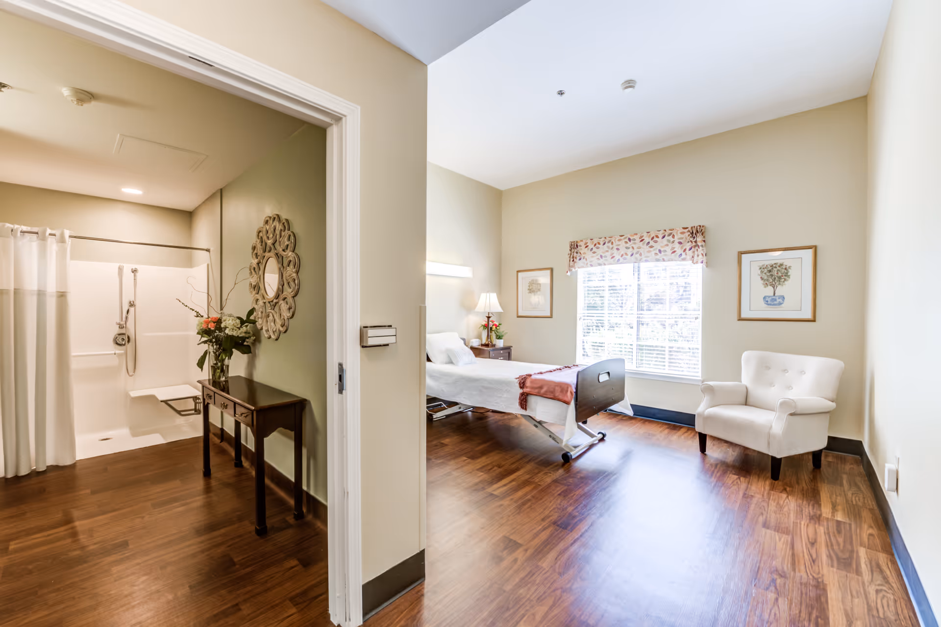 A bright senior living facility room with a hospital-style bed, a white armchair, and two framed pictures on the wall. The room has wooden flooring and a large window with blinds and a floral valance. To the left, there is an open doorway leading to a bathroom with a walk-in shower, a small table with a flower arrangement, and a decorative round mirror on the wall.