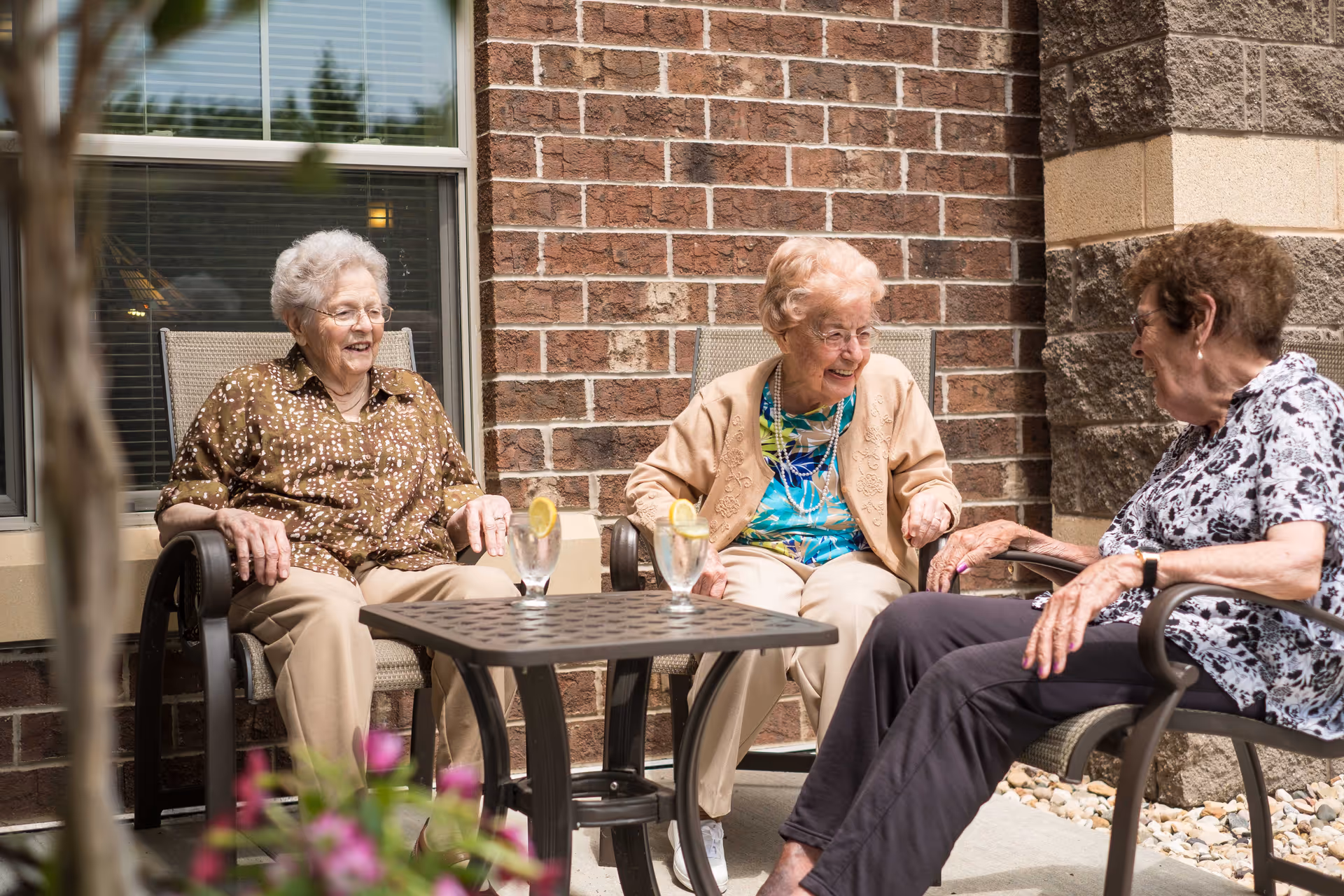 Three elderly women sitting outside on patio chairs around a small table with glasses of water and lemon, smiling and engaging in conversation. They are seated in front of a brick wall with a window and stone pillars visible.
