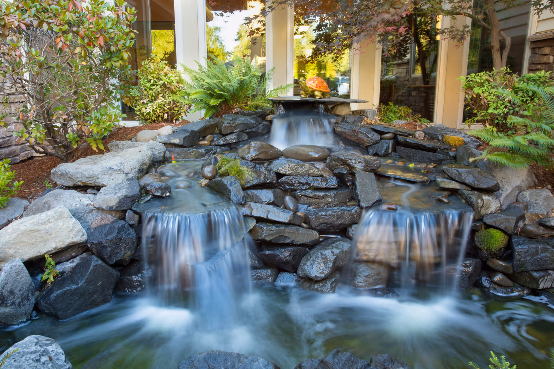 A tranquil outdoor water feature with cascading waterfalls flowing over rocks into a pond, surrounded by green plants and shrubs, with windows of a building visible in the background.