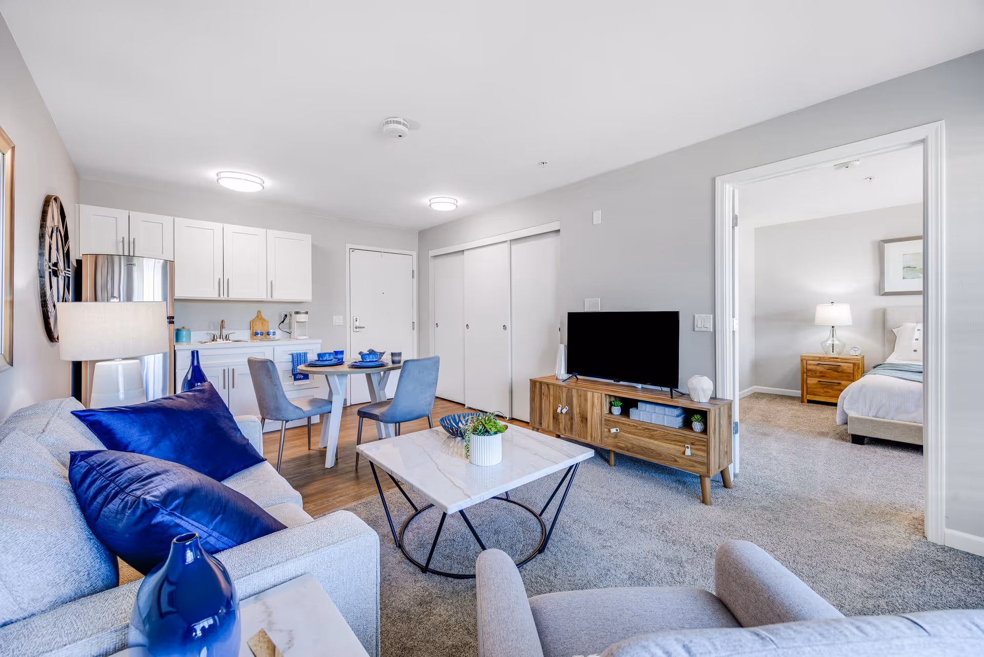 A modern living room and kitchen area in an apartment. The living room has a light gray sofa with dark blue pillows, a marble coffee table with a small plant, and a wooden TV stand with a flat-screen TV. The kitchen area features white cabinets, a small dining table set with blue dishes, and stainless steel appliances. A doorway leads to a bedroom with a bed, nightstand, and lamp.