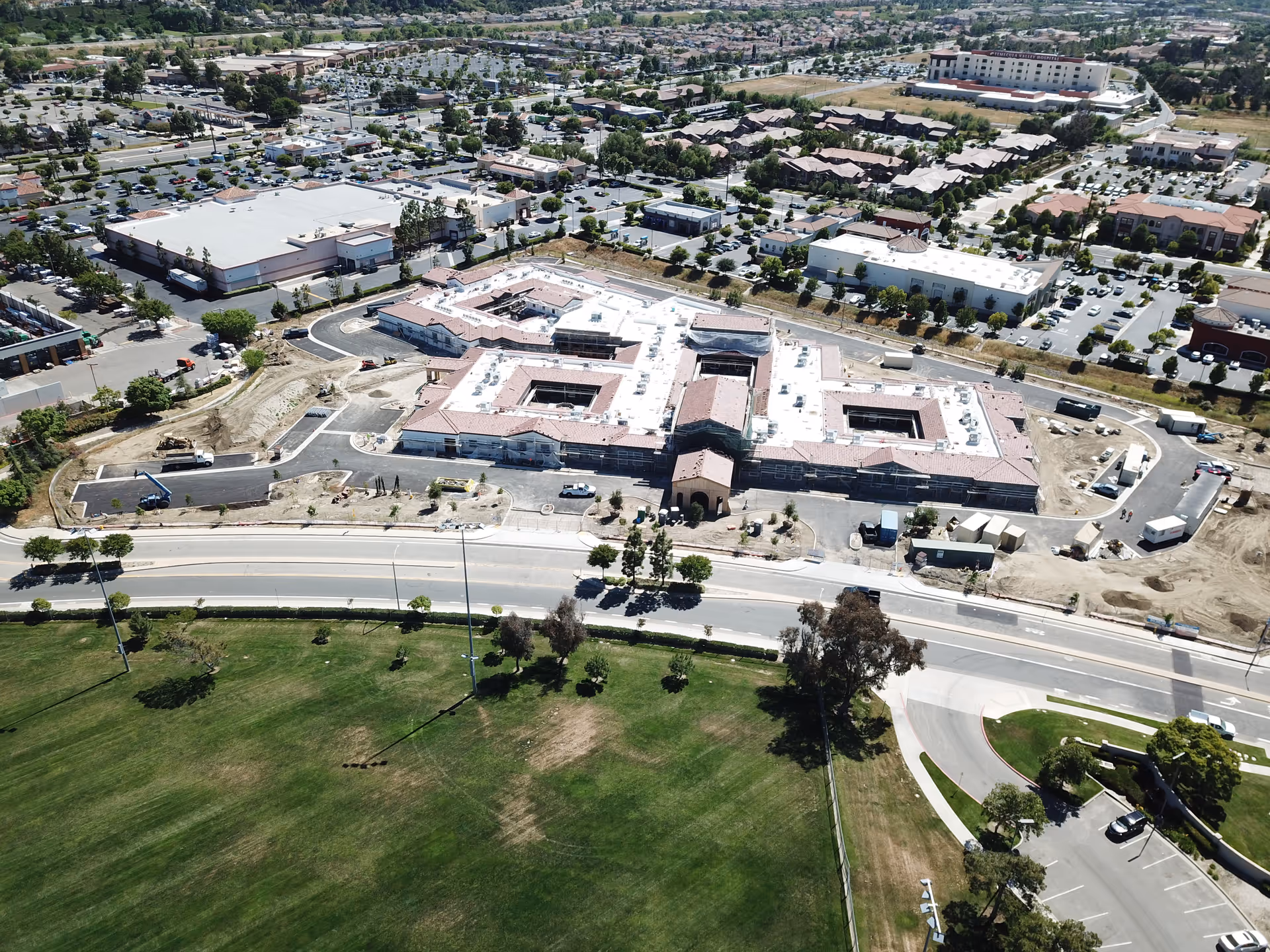 Aerial view of a large building complex under construction surrounded by roads, parking lots, and green spaces in a suburban area with many other buildings and houses in the background.