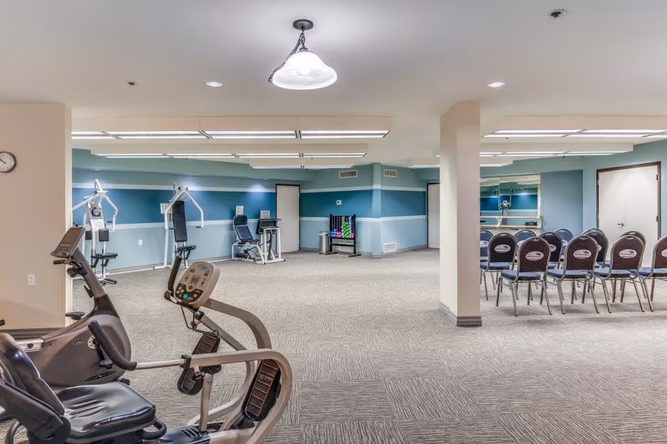 A spacious fitness and activity room in a senior living facility featuring exercise equipment such as stationary bikes and weight machines on the left side, and a row of chairs arranged for group activities or meetings on the right side. The room has light blue walls with a white stripe, carpeted flooring, and ceiling lights.
