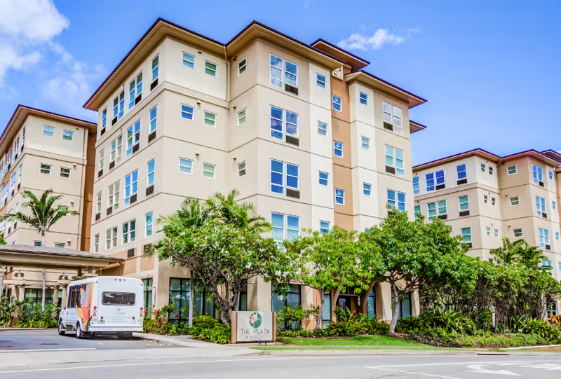 Three-story beige senior living building with green trees and a parked shuttle van in front.