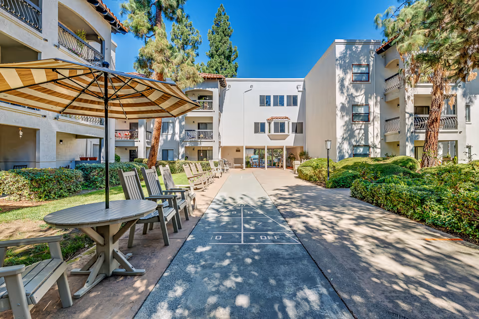 Outdoor courtyard area of a senior living facility with a shuffleboard court in the center, surrounded by chairs and tables with umbrellas. The courtyard is flanked by multi-story buildings with balconies, trees, and well-maintained bushes under a clear blue sky.
