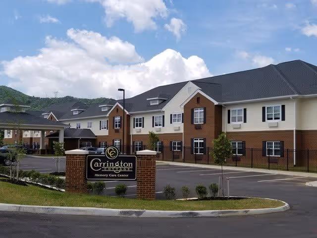 Exterior view of Carrington Cottage Memory Care Center, a two-story building with a combination of brick and light-colored siding. The building has multiple windows with black shutters and a dark roof. There is a parking lot in front and a sign displaying the facility's name near the entrance. The sky is partly cloudy with blue patches visible.