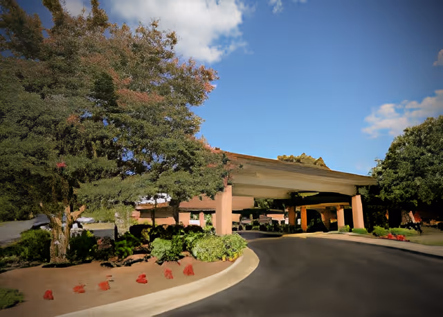 Entrance driveway of a nursing and rehabilitation facility with a covered drop-off area, surrounded by trees and landscaped greenery under a blue sky with some clouds.
