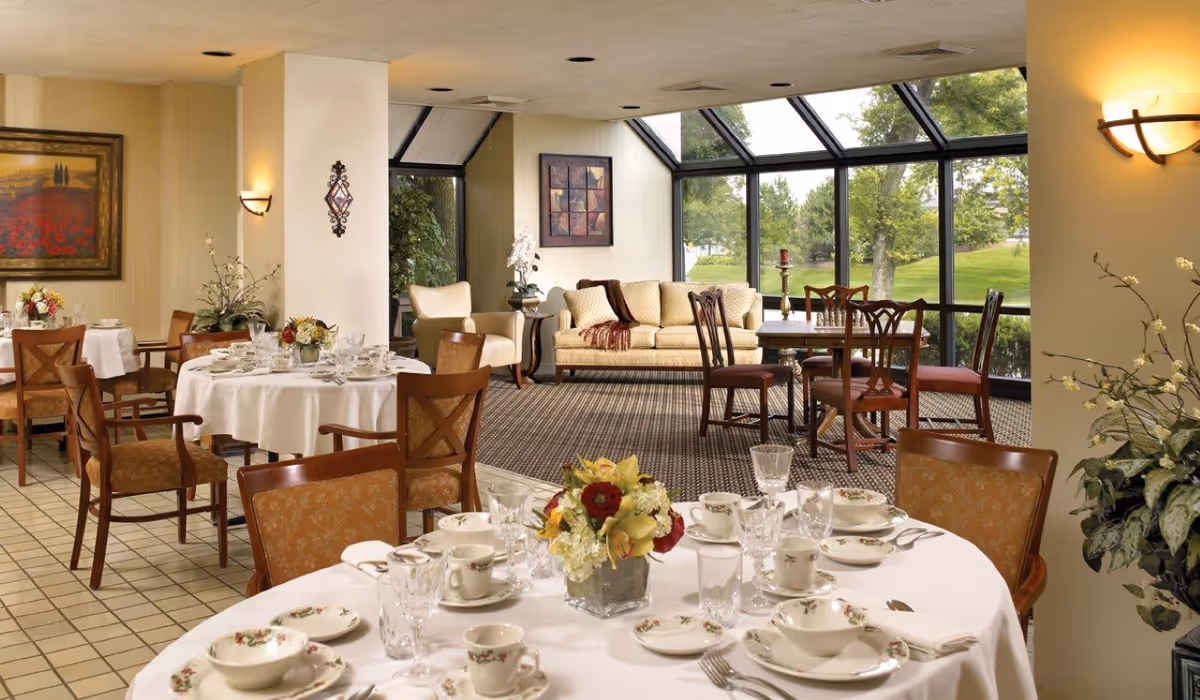 A bright dining area in a senior living facility with round tables set with floral-patterned dishes, glassware, and silverware. The room features wooden chairs with cushioned seats, a beige sofa, armchairs, and large windows providing a view of green trees and grass outside. Floral centerpieces decorate the tables, and framed artwork hangs on the walls.