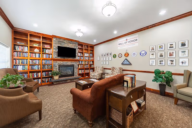 A cozy living room area in a senior living facility featuring a large built-in wooden bookshelf filled with books, a stone fireplace with a mounted flat-screen TV above it, several upholstered chairs and a sofa arranged around a coffee table, and a 'Wall of Honor' display with framed photos and military insignias on the wall.