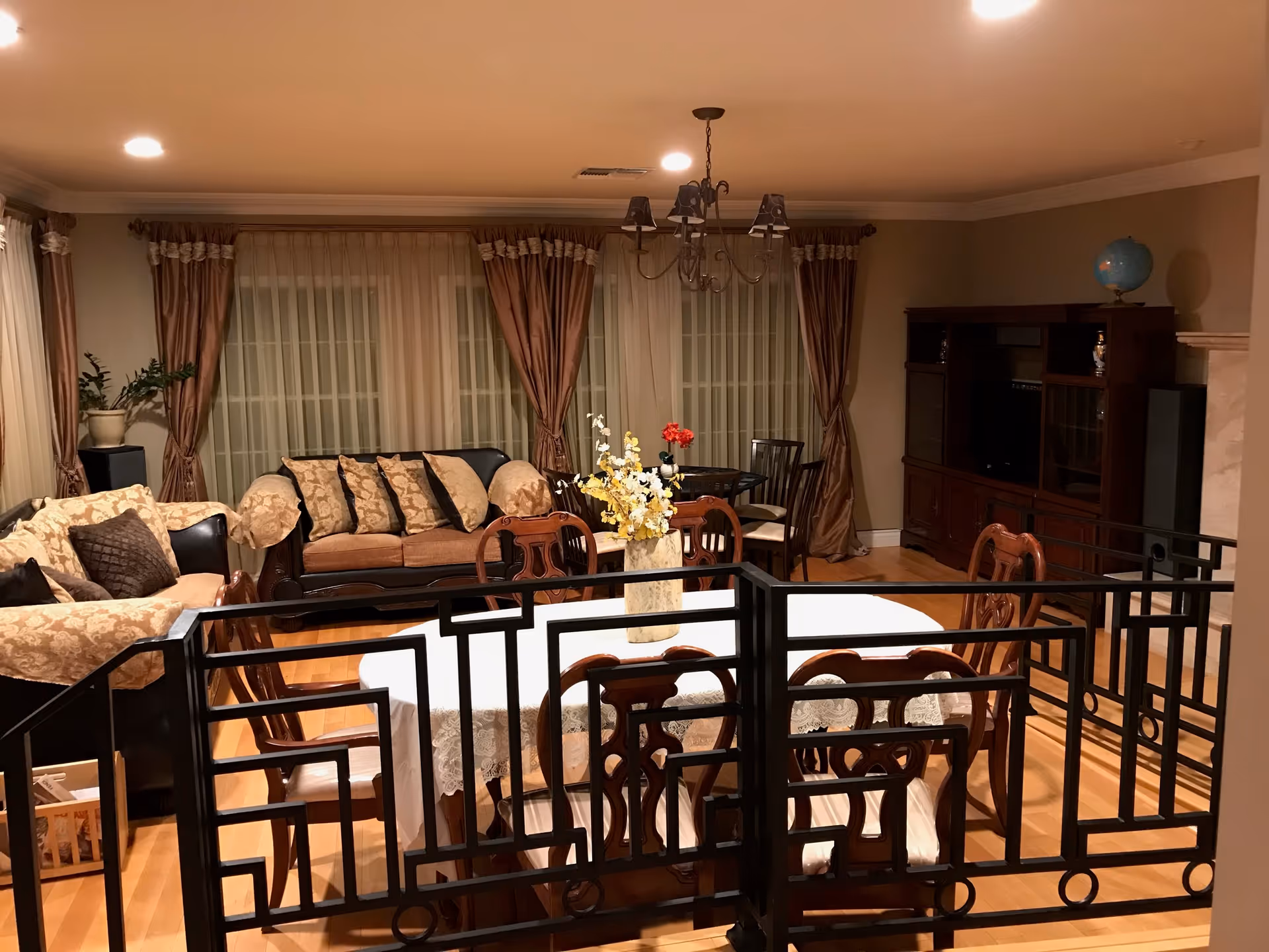 A cozy living room with a dining area featuring a round table covered with a white lace tablecloth and wooden chairs. Behind the dining area, there are two sofas with patterned cushions, large windows with sheer curtains and brown drapes, a wooden entertainment center with a globe on top, and a small round table with chairs and a vase of red flowers. The room has warm lighting and wooden flooring.