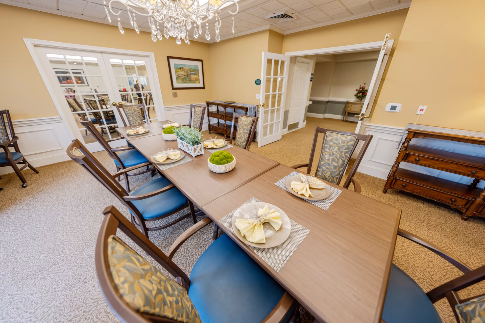 A dining room with a long wooden table set for six people with plates and folded napkins. The chairs have patterned backs and blue seats. There are decorative plants in the center of the table. The room has beige walls, white wainscoting, a chandelier, and double glass doors leading to another room.