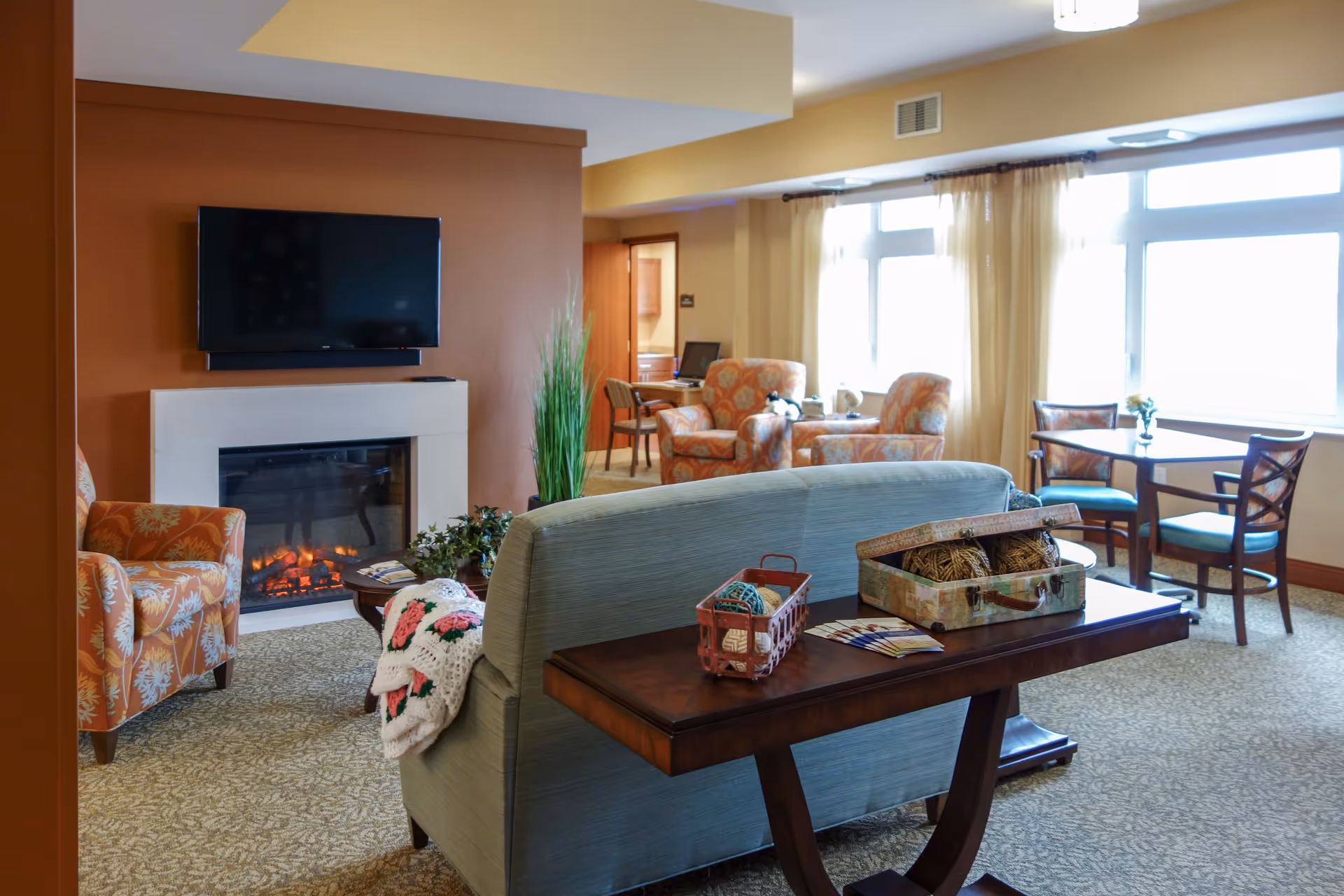 A cozy living room area in a senior living facility with a light blue sofa facing a wall-mounted flat screen TV above a modern fireplace. The room has patterned armchairs, a small table with yarn and knitting supplies, a desk with a computer, and large windows with sheer curtains letting in natural light.
