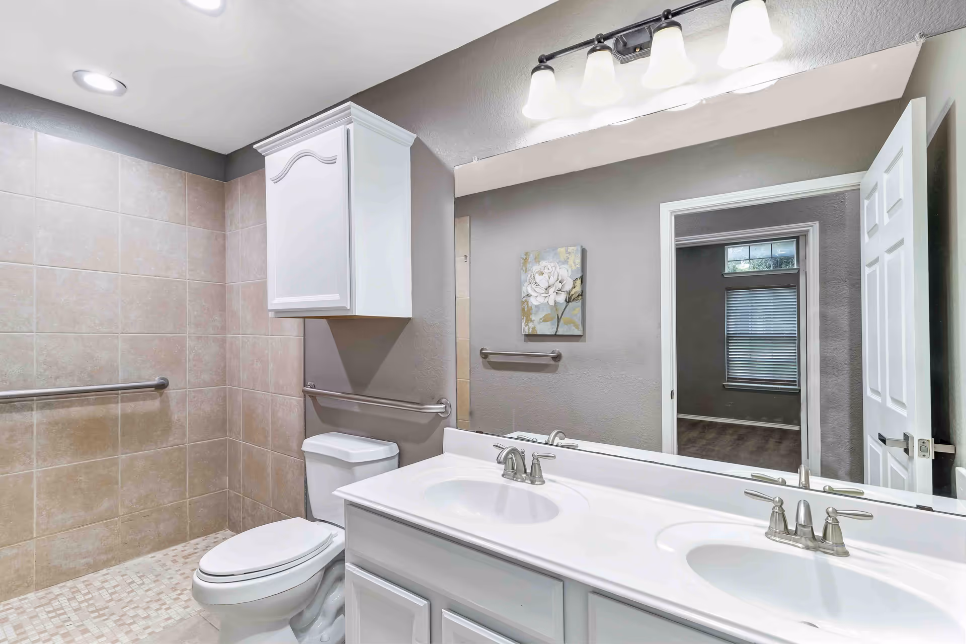 A clean and modern bathroom with beige tiled walls and floor. The bathroom features a white toilet, a white double sink vanity with silver faucets, a large mirror above the sinks, and a white cabinet mounted on the wall above the toilet. There are grab bars installed near the toilet and in the shower area. A door is open, showing a glimpse of an adjacent room with a window and blinds.