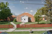 Single-story brick building with a centered white entryway, red picket fence, lawn and trees in front.
