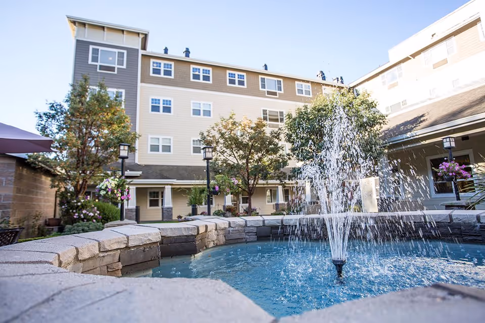 Outdoor courtyard area of a retirement community featuring a stone water fountain with water spraying upwards, surrounded by trees, flowers, and a multi-story building in the background under a clear blue sky.