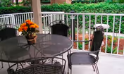 Outdoor patio area with a round metal table and four matching chairs. A small flower arrangement in a pot is placed on the table. The patio is surrounded by a white railing and greenery is visible in the background.