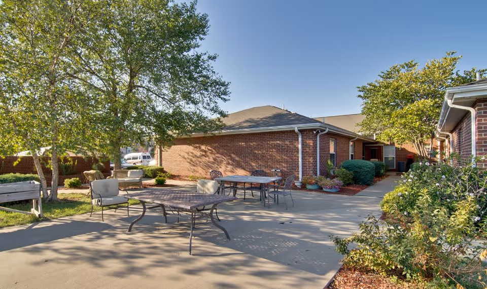 Outdoor patio area at a senior living facility with metal tables and chairs, surrounded by trees, bushes, and brick buildings under a clear blue sky.