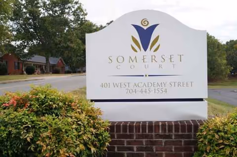 White Somerset Court sign with logo, brick base, and surrounding greenery.