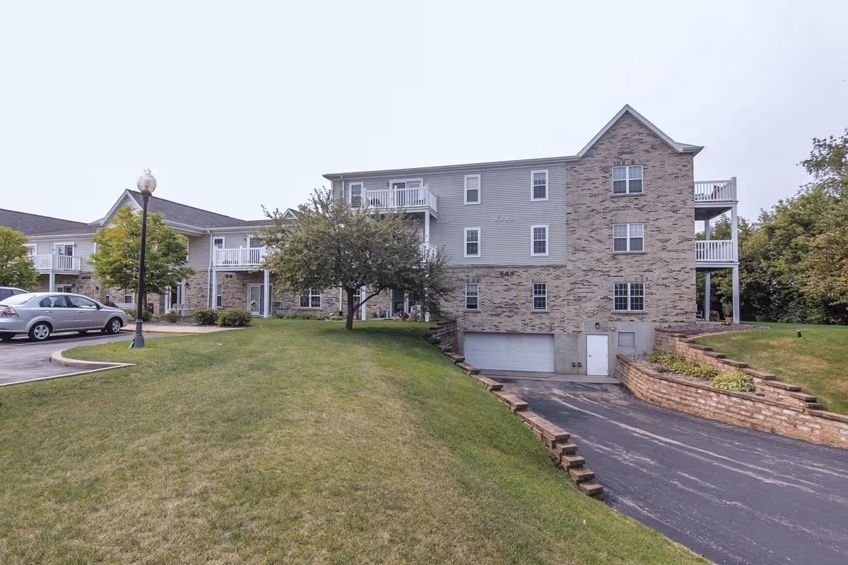 Exterior view of The Woodlands Senior Apartments showing a multi-story building with brick and siding facade, balconies, a driveway leading to a garage, a grassy lawn, a tree, a street lamp, and parked cars.