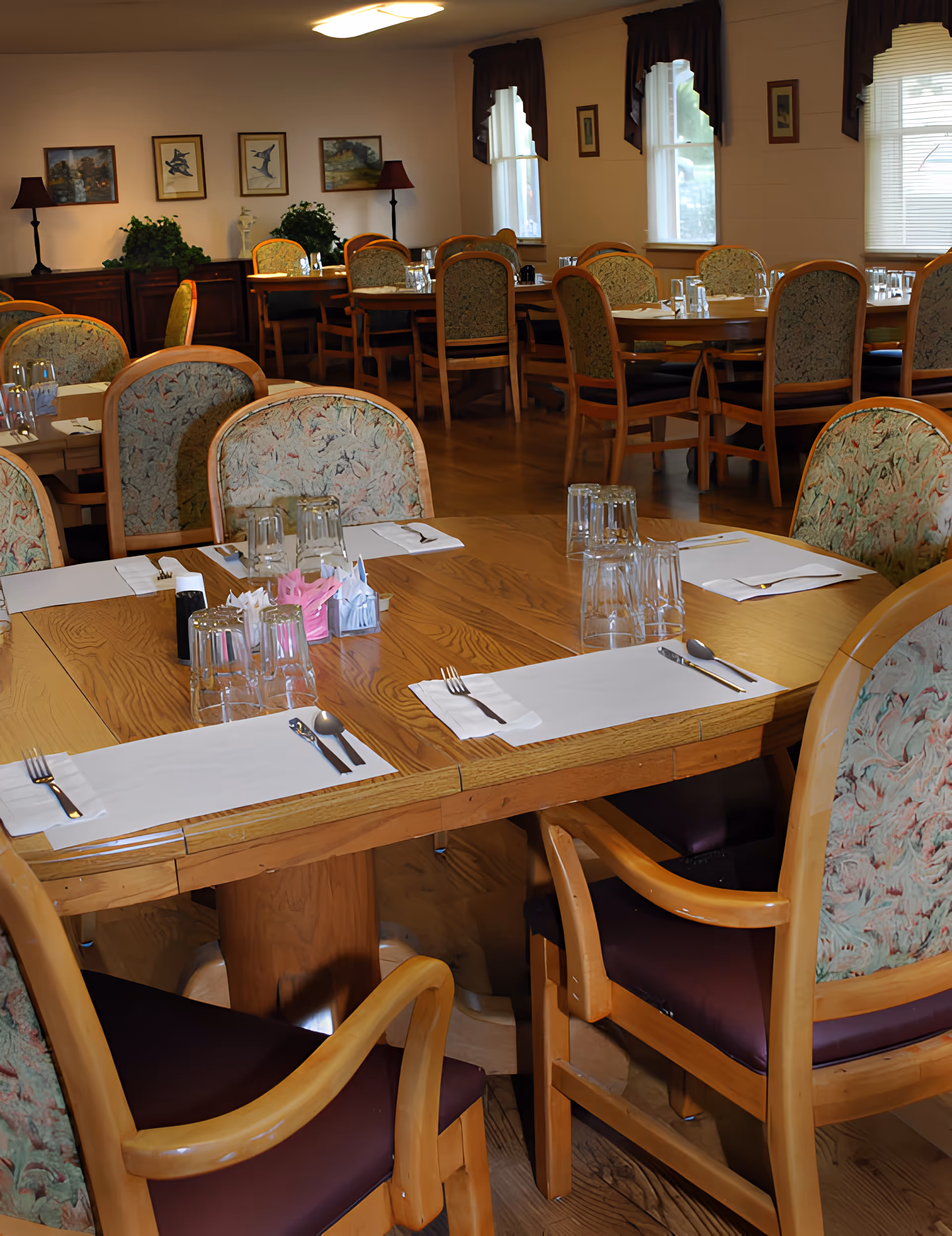 A dining room with multiple wooden tables and cushioned chairs arranged neatly. Each table is set with placemats, utensils, and upside-down drinking glasses. The room has wooden flooring, framed artwork on the walls, two floor lamps, and windows with valances allowing natural light to enter.