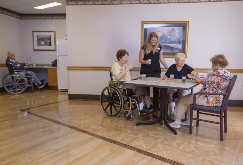 A caregiver stands with three elderly women seated around a table in a senior facility common/dining area while a man in a wheelchair sits near a small kitchenette in the background.