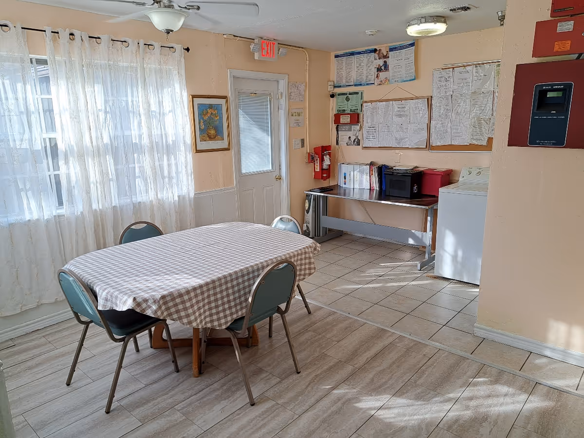 Bright communal dining room with a checkered-tablecloth table and chairs, bulletin boards, and a door under an exit sign.
