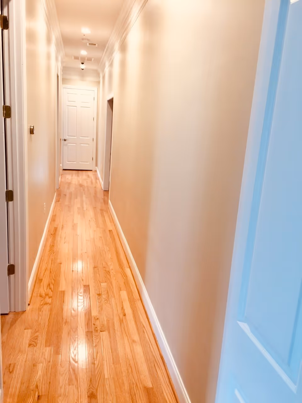 A long, narrow hallway with light beige walls and polished wooden flooring. There are several white doors along the hallway, with one door fully visible at the end. The hallway is well-lit with ceiling lights.