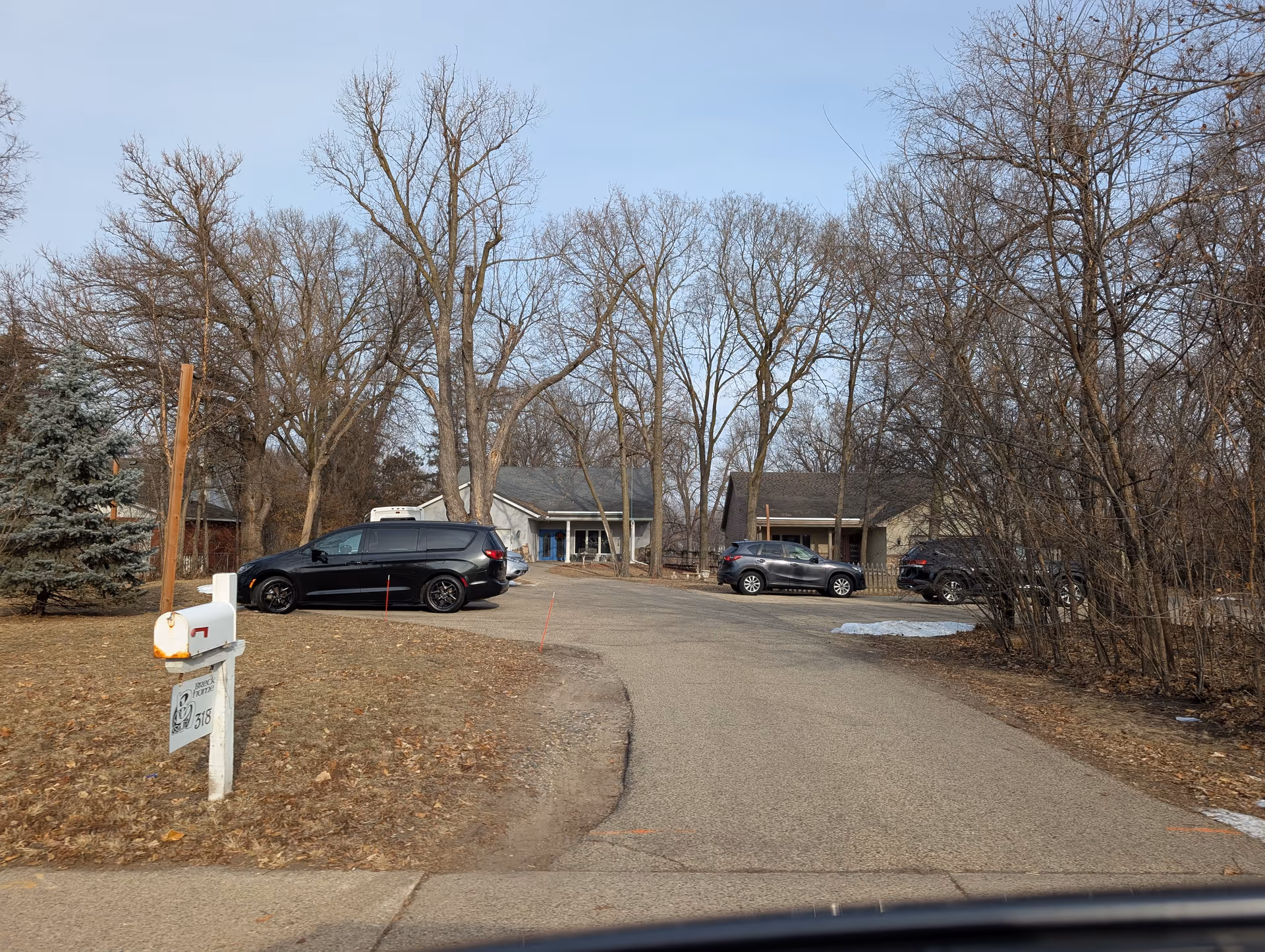 View of a residential care center driveway with three parked cars and two single-story houses surrounded by leafless trees in late autumn or early winter. A white mailbox with a sign reading 'Breck Home 318' is visible on the left side near the street.