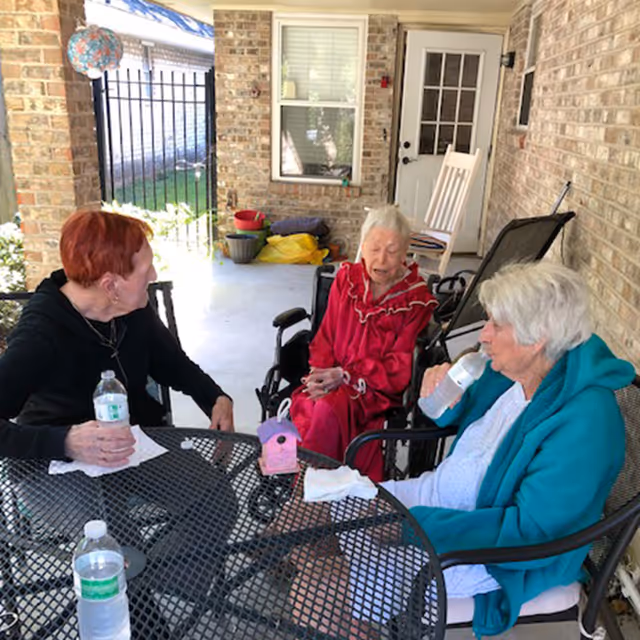 Three elderly women sitting and conversing on a covered patio area. One woman with red hair is holding a water bottle, another woman in a red dress is seated in a wheelchair, and the third woman with white hair is drinking from a water bottle. The patio has brick walls, a metal table with water bottles, and a rocking chair near a door in the background.