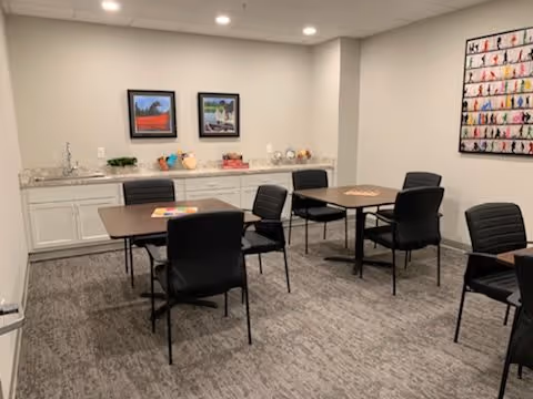 A small common area with four tables, each surrounded by black chairs, set on a carpeted floor. The back wall has white cabinets with decorative items on top, and two framed pictures hanging above. Another colorful framed artwork is visible on the right wall. The room is lit by recessed ceiling lights.