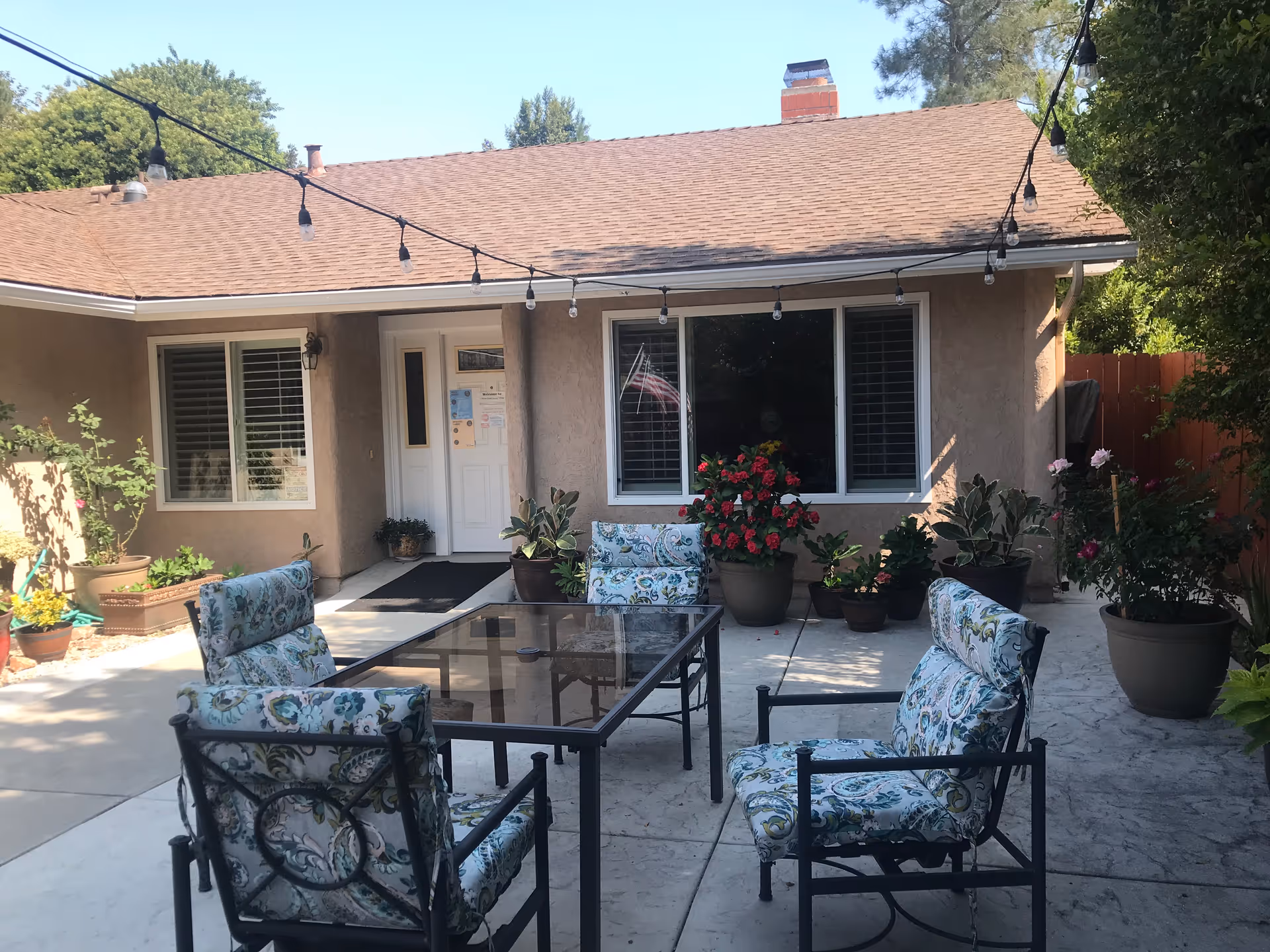 Outdoor patio area with a glass-top table surrounded by four cushioned chairs with floral patterns. The patio is adjacent to a beige building with two windows and a white door. Several potted plants and flowers are arranged around the patio, and string lights are hung overhead.