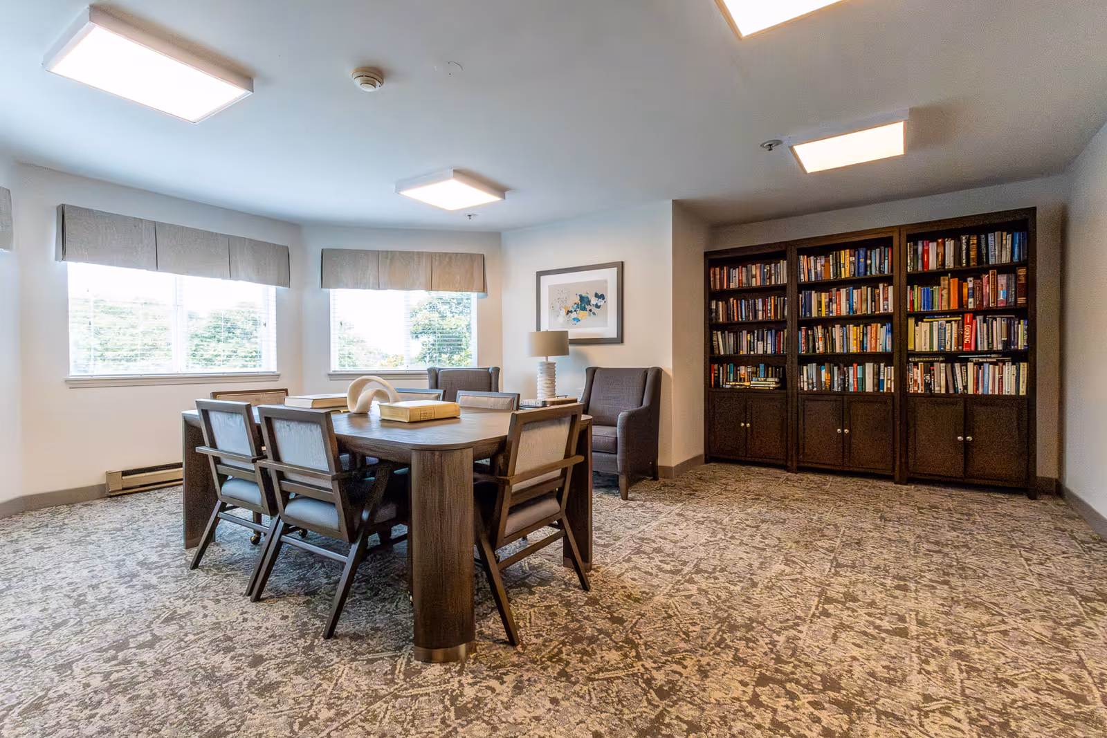 Well-lit communal room with a central wooden table and chairs, armchairs by the window, and bookshelves along one wall.