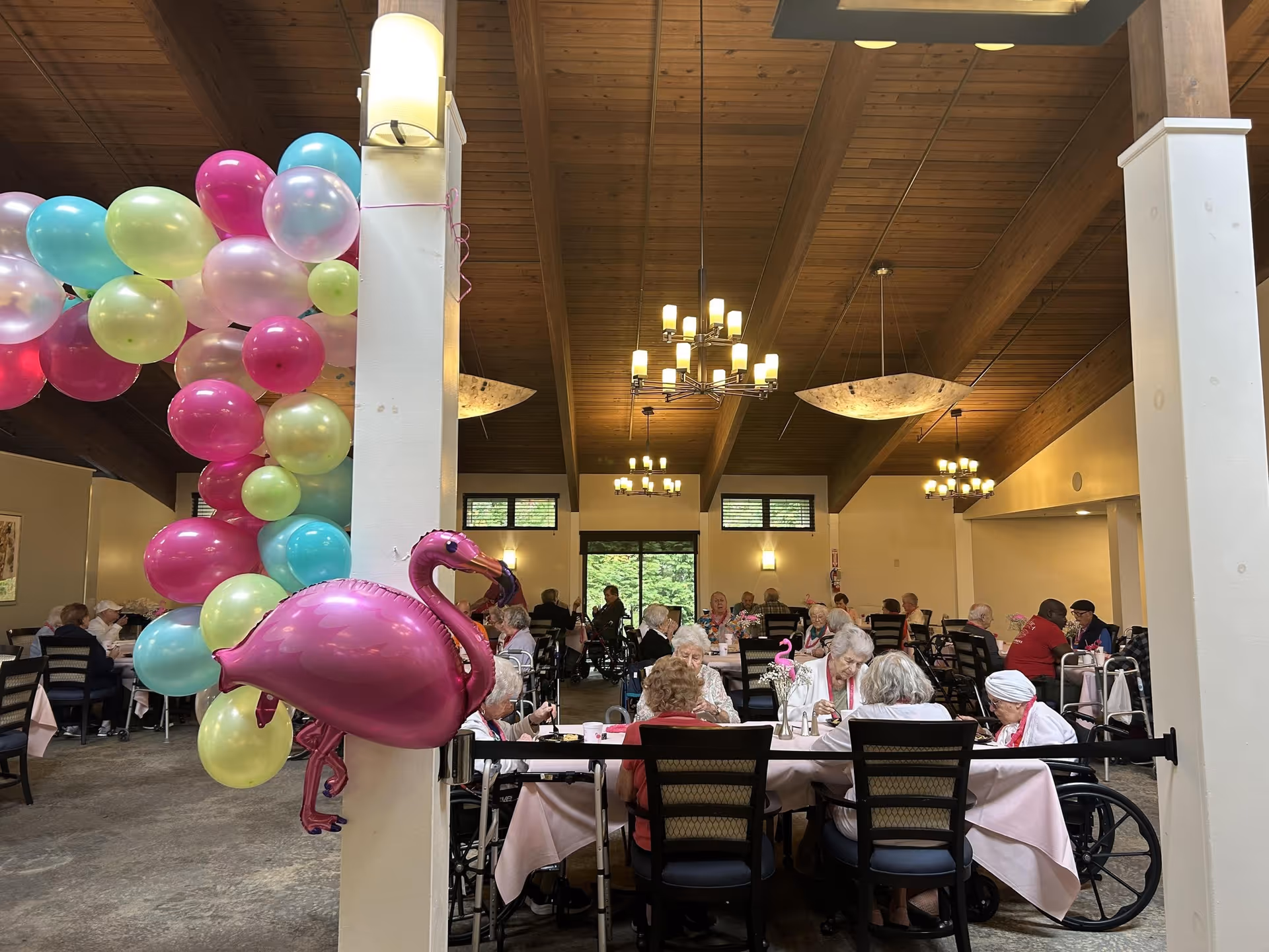 Large dining room at a senior living facility decorated with pastel balloons and a pink flamingo balloon, with residents seated at tables under wooden beams.
