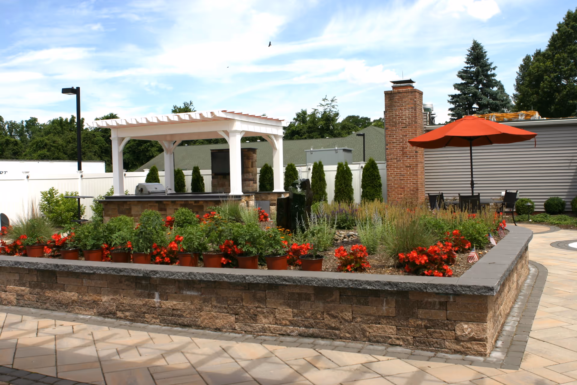 Outdoor garden area with a raised stone flower bed filled with red flowers and green plants. There is a white pergola structure with a built-in grill, a brick chimney, and a patio area with a table and chairs under a red umbrella. Trees and a building with gray siding are visible in the background under a partly cloudy sky.